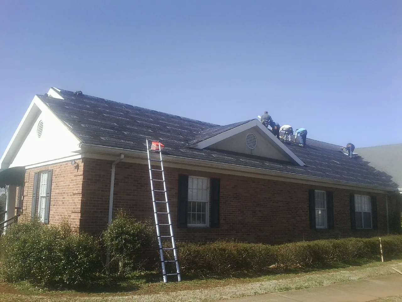 Crew tearing shingles off a brick ranch roof with a ladder set — Emergency Roof Repair work by our crew in Gambier