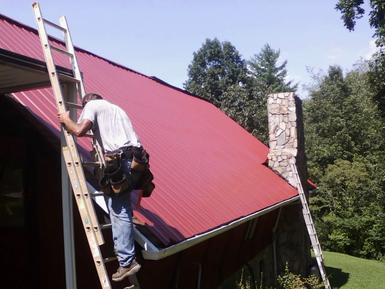 Roofer on a ladder beside a red metal roof and stone chimney — Soffit & Fascia Repair by Roofing Brothers in Gambier